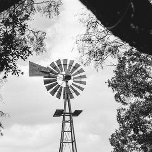 May include: A black and white photograph of a windmill with a metal frame and blades. The windmill is set against a cloudy sky and surrounded by trees.