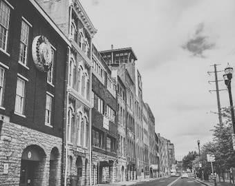 May include: A black and white photo of a city street with brick buildings. The buildings have many windows and some have decorative details. There are power lines running along the street.
