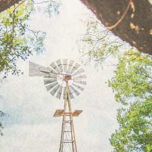 May include: A vintage windmill with a weathered metal blade structure against a cloudy sky. The windmill is surrounded by green trees.