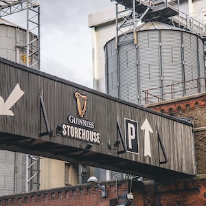 May include: A black and white sign with the words "Guinness Storehouse" and an arrow pointing up. The sign is attached to a building with large metal silos in the background.
