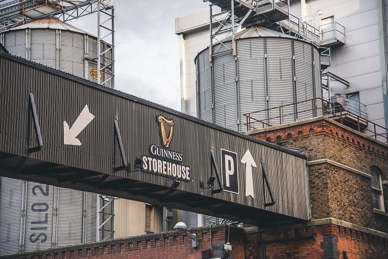 May include: A black and white sign with the words "Guinness Storehouse" and an arrow pointing up. The sign is attached to a building with large metal silos in the background.
