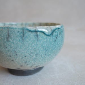 Op de afbeelding: Een handgemaakte keramische kom met een uniek, gekraakt turquoise glazuur. De kom heeft een donkergrijze basis en een zacht golvende rand. De textuur van het glazuur is zichtbaar, wat bijdraagt aan de ambachtelijke kwaliteit.