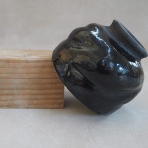 Pode incluir: Uma escultura de cerâmica preta com acabamento brilhante. A forma abstrata tem um corpo arredondado e uma abertura alargada. Um pequeno bloco de madeira retangular está ao lado da escultura.