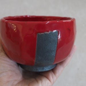 Può includere: Una ciotola in ceramica fatta a mano con un esterno rosso lucido e un accento verticale grigio scuro. La ciotola ha una forma arrotondata e una piccola base grigio scuro. La ciotola è tenuta in una mano.
