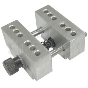 Op de afbeelding: Een zilveren metalen klem met een veer en een knop. De klem heeft meerdere gaten aan elke kant.