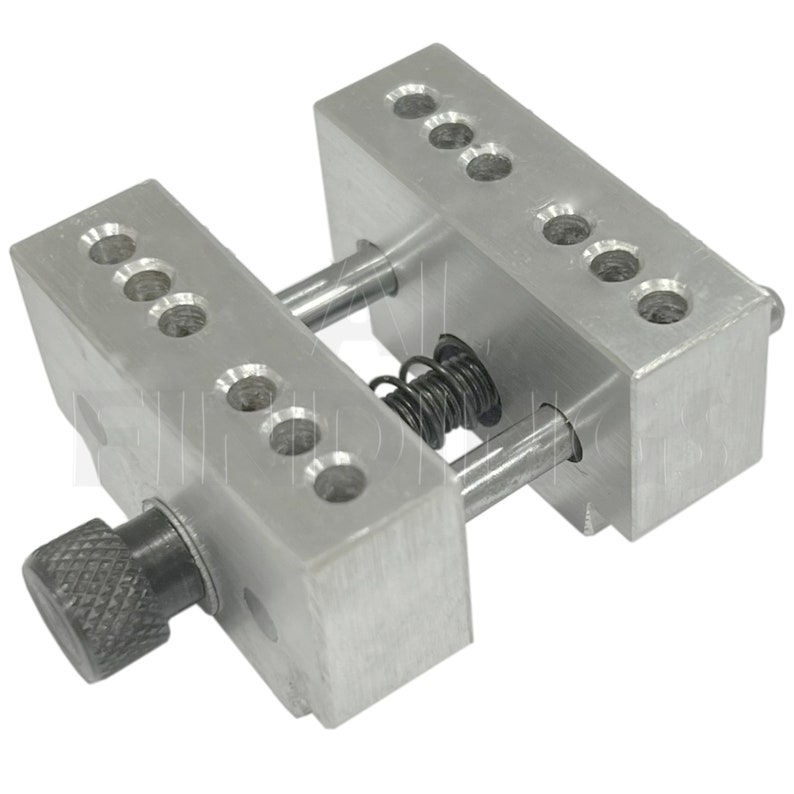 Op de afbeelding: Een zilveren metalen klem met een veer en een knop. De klem heeft meerdere gaten aan elke kant.