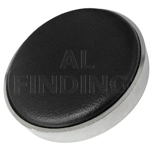 Puede incluir: Un cojín de reloj de cuero negro con un borde de metal plateado. El cojín tiene el texto "AL FINDING" grabado en relieve.