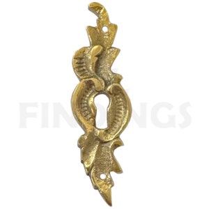 Puede incluir: Una tapa de mirilla de metal dorado con un diseño decorativo y ornamentado. La tapa tiene una mirilla en el centro y dos pequeños agujeros a cada lado.