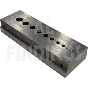 May include: A rectangular, gray metal tool with a series of graduated holes and a central groove. The tool appears to be made of solid metal and is designed for a specific purpose, likely in a workshop or craft setting.
