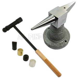 Puede incluir: Un yunque de metal plateado para hacer joyas con una parte superior puntiaguda y una base redonda. El yunque está sobre una base gris. Un martillo negro con un mango de madera y una cabeza redonda está al lado del yunque. Hay cuatro pequeñas herramientas de metal y plástico al lado del martillo.