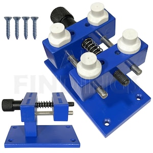 May include: A blue metal watchmaker's vise with a spring-loaded clamp and four white plastic jaws. The vise is mounted on a blue metal base with four screw holes. There are four screws included.