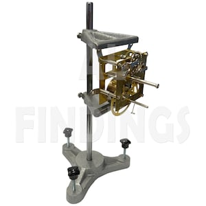 May include: A gray metal clock movement holder with a three-legged base and adjustable arms. The holder supports a brass clock movement with visible gears and components. The word "FINDINGS" is partially visible in the background.
