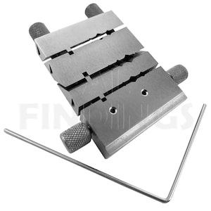 Op de afbeelding: Een metalen ringmandrel met vier sleuven voor het bepalen van de ringmaat. De mandrel heeft aan elke kant verstelbare knoppen en een kleine metalen staaf inbegrepen.