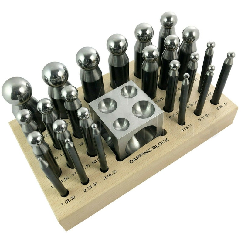 Op de afbeelding: Een houten dappingblok met een set metalen dappingponsen en een metalen dappingblok met vier verschillende inkepingen. De dappingponsen zijn gerangschikt in een raster op het houten blok en gelabeld met hun afmetingen in inches. Het dappingblok is gelabeld met "DAPPING BLOCK".