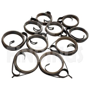 Puede incluir: Un conjunto de nueve resortes helicoidales de metal oscuro dispuestos en grupo. Cada resorte está formado por una tira de metal fina y plana enrollada en forma circular con un pequeño bucle que sobresale en un extremo. El metal tiene un acabado oscuro.