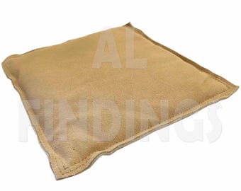 May include: A square, tan-colored bean bag. The bean bag is made of a soft, suede-like material and has a stitched border. The text "AL FINDINGS" is partially visible on the surface of the bean bag.