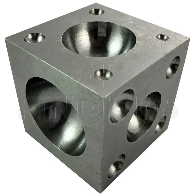 Op de afbeelding: Een metalen kubus met vier verschillende afgeronde inkepingen aan elke kant. De kubus is gemaakt van zilverkleurig metaal en heeft een gladde, gepolijste afwerking.