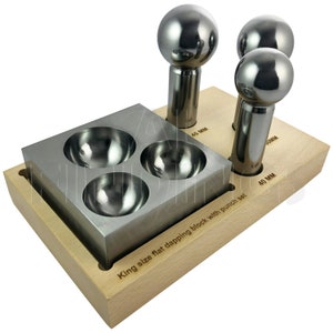 Puede incluir: Un bloque de madera con tres punzones de dapping de metal y un bloque de dapping de metal con tres depresiones redondas. El bloque tiene el texto "King size flat dapping block with punch set" grabado. Los punzones están etiquetados con 40 mm, 45 mm y 50 mm.