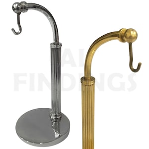 Puede incluir: Dos expositores metálicos. Uno es de color plateado con una base redonda y un gancho curvo. El otro es de color dorado con un diseño similar. Ambos soportes tienen una columna estriada y están diseñados para sostener objetos.