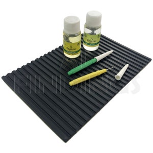 May include: Two small bottles of clock oil and watch oil with white caps and green labels. The bottles are on a black rubber mat with raised ridges. There are two small tools with white tips and green and yellow handles.