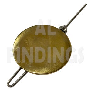 May include: A circular brass clock pendulum bob with a brushed metallic finish. The component features a thin, straight metal rod extending from the top, secured by a knurled adjustment nut, and a U-shaped wire loop extending from the bottom edge.