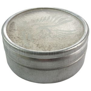 May include: A small, round, silver-colored tin filled with a light-colored powder. The lid has a frosted design of a stylized bird or dragon. The tin is sitting on a white background.