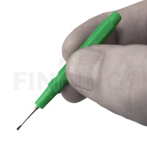 May include: A green plastic tool with a pointed metal tip. The tool is held in a gloved hand.