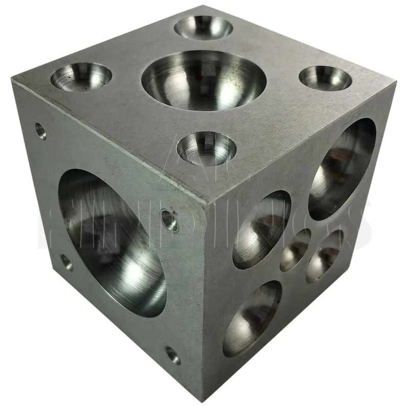 Op de afbeelding: Een metalen kubus met zes ronde inkepingen van verschillende grootte. De kubus is gemaakt van zilverkleurig metaal en heeft een gladde, gepolijste afwerking.
