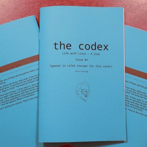 May include: The cover of a blue zine titled "the codex" with the subtitle "Life with Linux - A Zine" and the issue number "Issue #3". The text "Typeset in LaTeX (except for this cover)" is also included. The cover features a simple line drawing of a person wearing glasses.