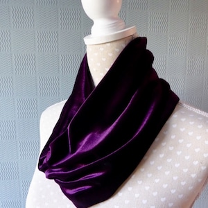 May include: A deep purple velvet infinity scarf draped over a mannequin. The scarf is a rich, solid color, with a soft, lustrous texture. The mannequin is white with a cream-colored fabric with white hearts.