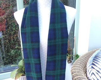 May include: A blue and green plaid scarf with fringe at the ends is displayed on a mannequin. The scarf is a classic tartan pattern, with dark blue and green intersecting lines forming a grid. The scarf is made of a soft material.