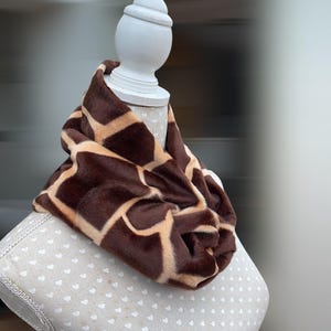 May include: A brown and tan giraffe print infinity scarf displayed on a mannequin. The faux fur scarf has a soft, plush texture and a repeating pattern of brown and tan patches. A cosy accessory.
