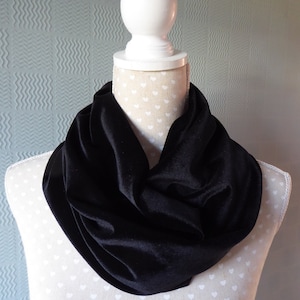 May include: A black velvet infinity scarf draped on a mannequin. The scarf is a deep, solid black and has a soft, luxurious texture. The mannequin is white with a heart pattern, and the background is a light blue.