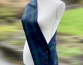 May include: A blue and green plaid sash draped over a mannequin, secured with a gold safety pin. The fabric has a classic tartan pattern. The mannequin is white with a gray head and is set against a blurred outdoor background.