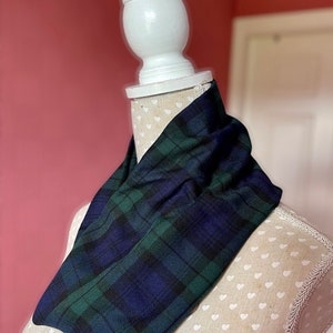 May include: A dark blue and green tartan infinity scarf is displayed on a mannequin. The scarf's pattern is a classic plaid design. The mannequin is set against a pink backdrop.