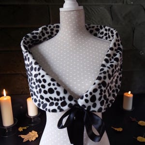 May include: A white faux fur shawl featuring a black dalmatian-style spotted pattern. The shawl is draped over a mannequin and secured at the front with a black satin ribbon tied into a bow. The fabric has a soft, plush texture throughout.