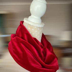 May include: A vibrant red velvet infinity scarf draped on a white mannequin head. The scarf is soft and flowing, with a luxurious sheen. The mannequin head is on a blurred background.