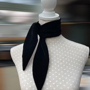 May include: A black velvet neck scarf is draped around a mannequin. The scarf is tied in a loose knot, with long, pointed ends. The mannequin is white with a heart-patterned fabric. The background is blurred with shades of grey and white.