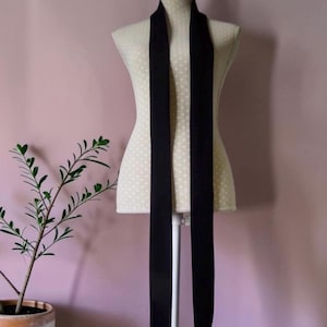 May include: A long, narrow black scarf draped vertically over a cream-coloured mannequin torso. The scarf features a smooth, solid black fabric that hangs straight down from the neck area, creating two parallel, elongated ends.