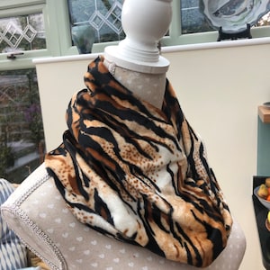 May include: A faux fur infinity scarf with a tiger stripe pattern in shades of brown, black, and cream. The scarf is draped over a mannequin, showcasing its circular design and soft texture. This accessory adds a touch of animal print to any outfit.