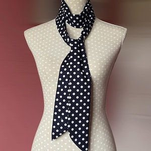Könnte beinhalten: Ein marineblauer Schal mit weißen Polka-Dots ist auf einer Schaufensterpuppe drapiert. Der Schal ist geknotet, wobei die Enden herunterhängen. Die Schaufensterpuppe ist beige mit einem weißen Herzmuster. Der Hintergrund ist ein verschwommenes Rosa.