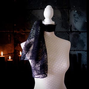 May include: A black scarf featuring a detailed spiderweb pattern draped around the neck of a white mannequin. The scarf is made of a sheer, lightweight fabric with a distinct web-like design that covers the entire surface of the accessory.