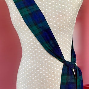 Puede incluir: Una faja a cuadros azules y verdes está drapeada sobre un maniquí. La faja está anudada a la cintura. El maniquí está cubierto con una tela color crema con un patrón de pequeños corazones blancos.