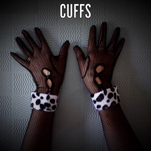 Pode incluir: Luvas de malha pretas com detalhes recortados e punho branco e preto com manchas. A palavra "CUFFS" é exibida em texto branco na parte superior da imagem. As luvas são usadas em uma superfície texturizada azul clara.