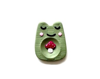May include: A green frog-shaped trinket dish with a red and white mushroom in the center. The frog has closed eyes, pink cheeks, and a smiling mouth. The dish has a textured surface.