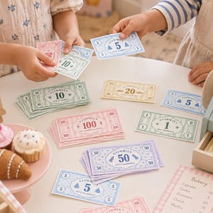 May include: A toy cash register with a paper receipt reading Good Things Ahead, stacks of colorful Glimmies play money in denominations of 1, 5, 10, 20, 50, and 100, a small wooden sign reading Fresh Treats Always a Good Idea, and a bakery menu.