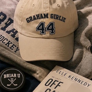 Puede incluir: Una gorra de béisbol beige con el texto bordado GRAHAM GIRLIE y el número 44 en azul oscuro. La gorra está colocada junto a un libro titulado OFF CAMPUS de Elle Kennedy, un disco de hockey negro con el texto BRIAR U y una sudadera gris con capucha.