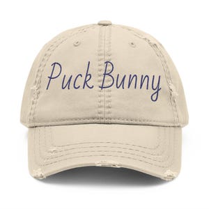May include: A beige distressed baseball cap featuring the text Puck Bunny embroidered in dark blue cursive script across the front panels. The hat has a curved brim with frayed edges and a six-panel construction with matching top button.