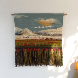 Puede incluir: Tapiz de pared tejido a mano que representa un paisaje con cielo azul, nubes blancas y amarillas, y un campo verde y rojo. El borde inferior presenta largas borlas con flecos en tonos marr&oacute;n, amarillo y rojo. El tapiz est&aacute; montado en una barra de madera.