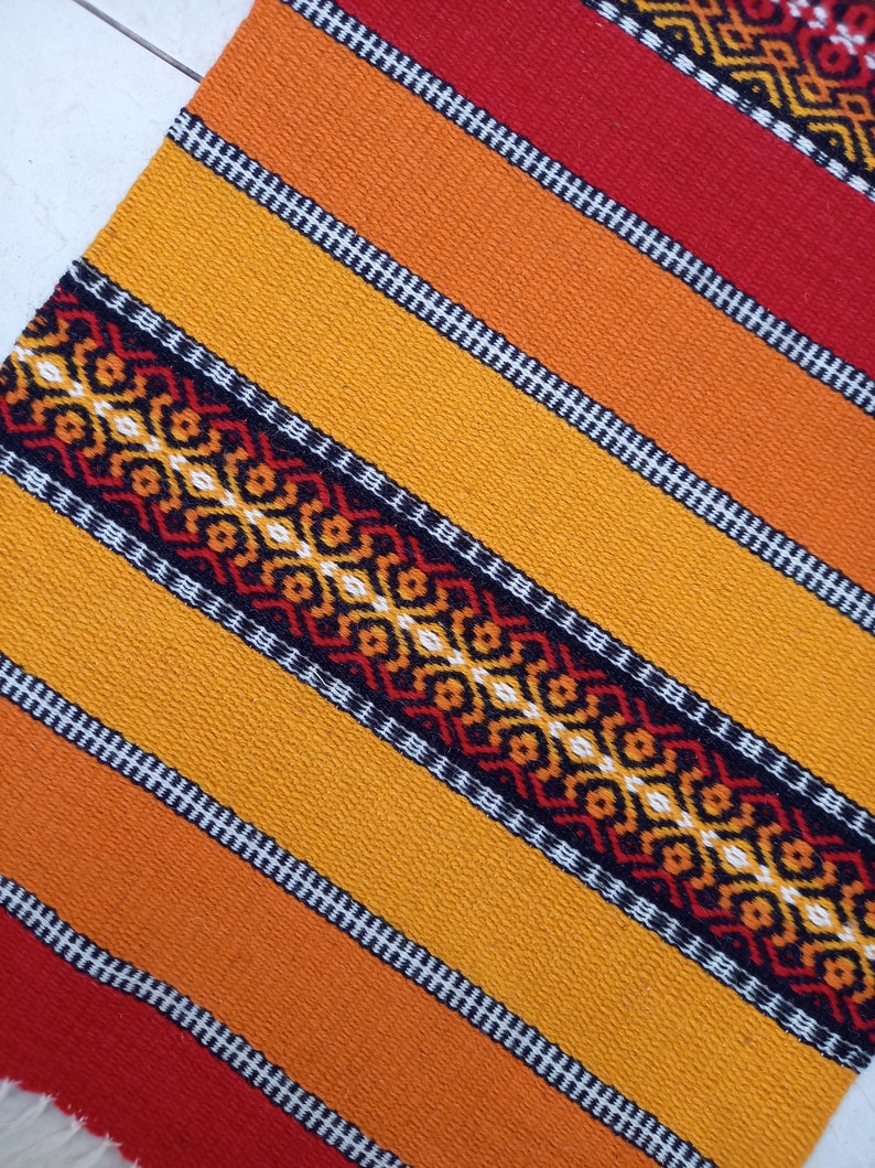 May include: A colorful woven table runner with a geometric pattern in shades of red, orange, yellow, and black.